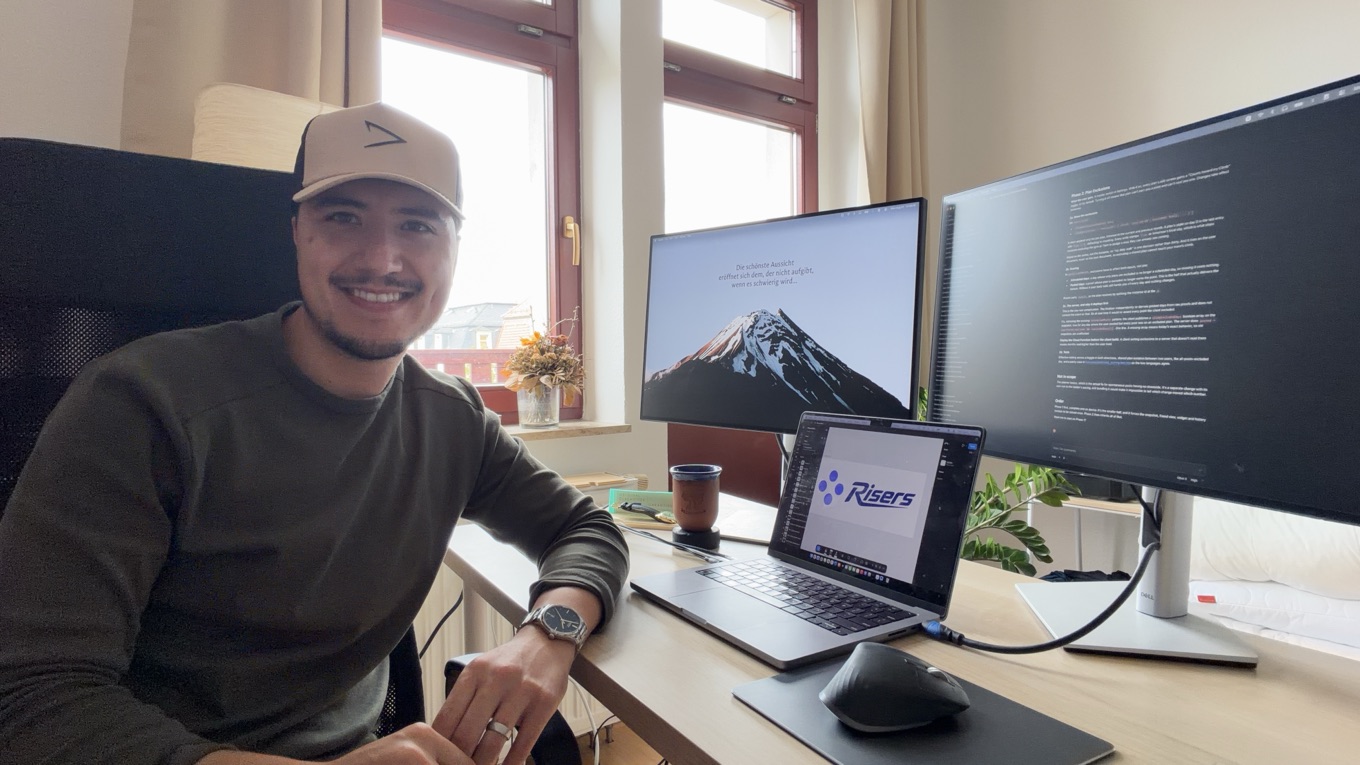 Dylan at his desk, smiling, with the Risers logo open on a laptop and code on the monitor behind him.